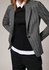 Blazer gris en chevrons à coupe ajustée, associé à un pull noir côtelé et une chemise blanche à col, présentant une texture lisse et un subtil effet de superposition.