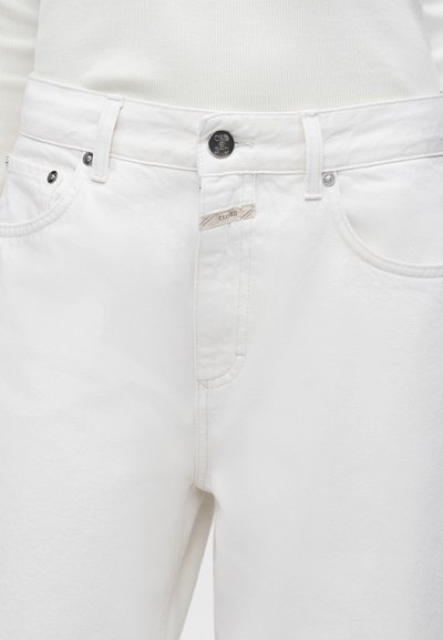Gros plan d'un jean en denim blanc à l'avant avec un bouton argenté et une étiquette de marque visible sur une petite étiquette en tissu près de la fermeture éclair.