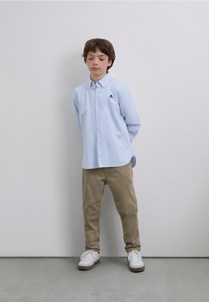 Camisa de botones a rayas azul claro con un pequeño logo en el pecho, combinada con pantalones chinos beige y zapatillas blancas, de pie frente a una pared lisa.