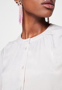 Blusa rosa chiaro con scollo rotondo e chiusura frontale a bottoni. Presenta pieghe sottili e un tessuto testurizzato. Orecchini a chandelier di grandi dimensioni completano il look.