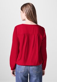 Rode blouse met lange mouwen en een geraffineerde achterkant, ronde halslijn en elastische manchetten, gecombineerd met lichtblauwe hoog-getailleerde jeans.