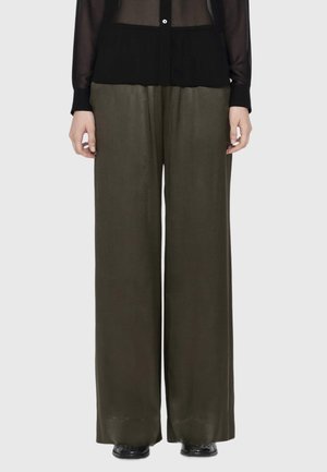 Person wearing olive green wide-leg pants, black sheer buttoned top, and black shoes, standing against a plain light background.