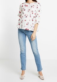 Blusa floreale bianca con fiori rossi e teal, maniche 3/4, abbinata a jeans skinny azzurri e scarpe bianche con tacco largo e traforato.