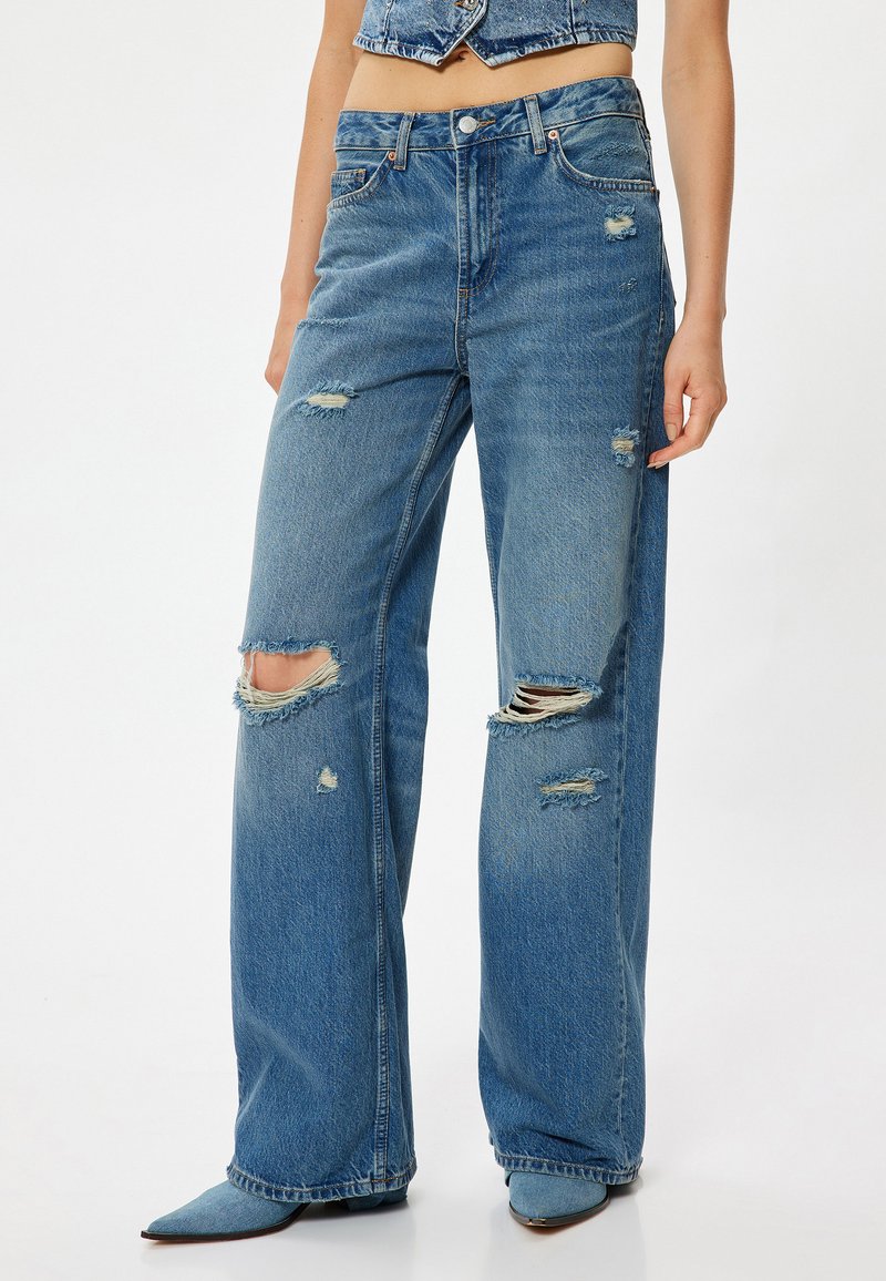 Jeans de pierna ancha de un azul claro desgastado con grandes rasgaduras en las rodillas, de cintura alta y con un sutil desvanecimiento, fabricados en tela de mezclilla con un acabado ligeramente texturizado.