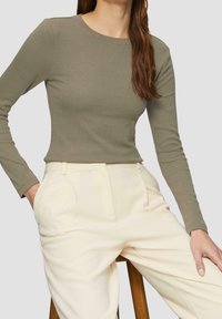 Top de manga larga de color verde oliva con textura de canalé, combinado con pantalones crema ajustados, con bolsillos laterales y un corte relajado, sentado en un taburete de madera.