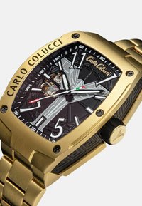 Gouden roestvrijstalen horloge met een zwarte gestructureerde wijzerplaat, zilveren accenten, zichtbare beweging en een rubberen band. Bevat gedurfde cijfers.