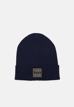 Navy blue knitted beanie with a folded cuff, featuring a gray fabric patch with embossed logo. Smooth texture and rounded top design.