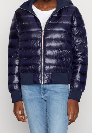 Personne portant une doudoune bleu marine brillante avec fermeture éclair, poignets et ourlet côtelés, par-dessus une chemise blanche et un jean bleu clair.
