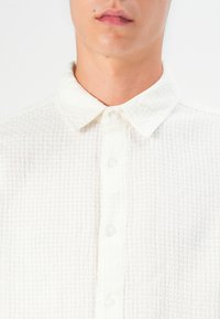Camisa de manga corta, blanca, con una tela texturizada de tejido en waffles, que presenta un cuello con botones y botones en la parte frontal.