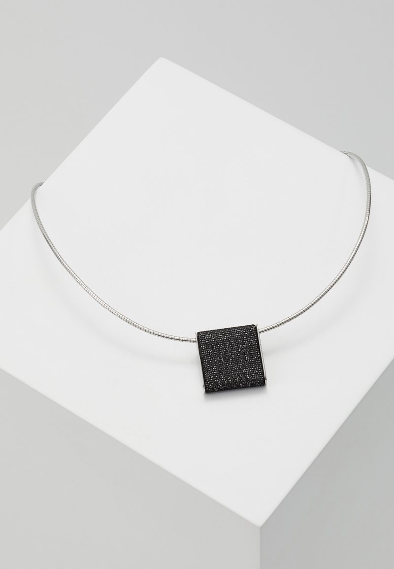Collier en métal argenté avec un pendentif carré noir texturé présenté sur une surface blanche inclinée.