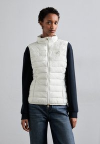 Bijela puffer prsluk s visokim ovratnikom i zatvaranjem na zip, uparen s tamnim dizajnom rukava. Upadljiv zlatni zip i reljefni detalj s logotipom.