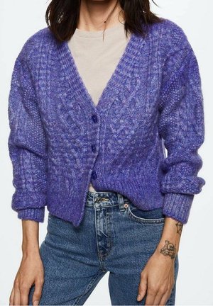 Kobieta ubrana w fioletowy kardigan w warkocze na beżowej koszuli i niebieskich dżinsach, z tatuażem czaszki i skrzyżowanych kości na prawym nadgarstku.