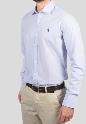 Homme portant une chemise bleu clair à fines rayures rentrée dans un pantalon beige avec une ceinture marron foncé sur un fond uni.