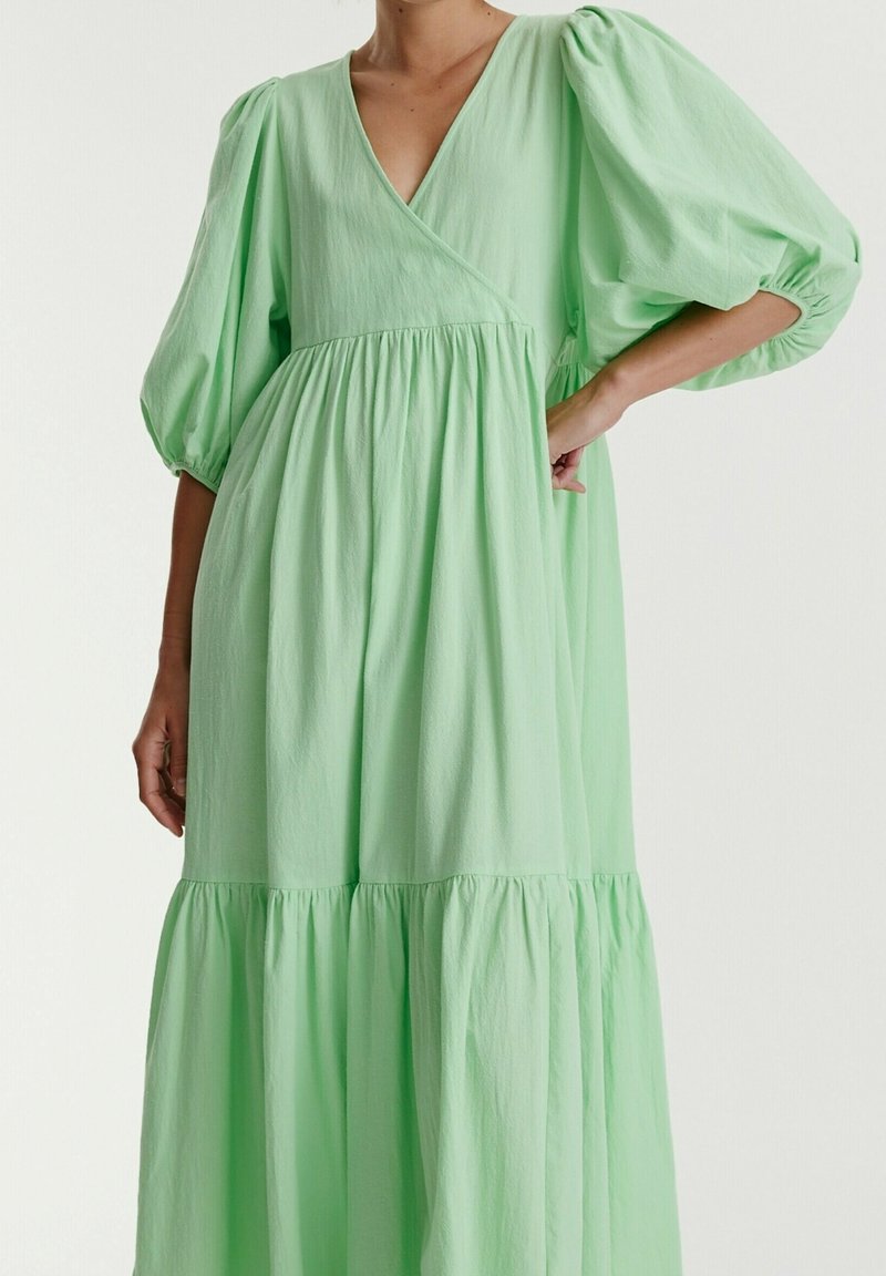 Femme portant une robe ample vert clair avec des manches trois-quarts bouffantes et une jupe à volants froncés, debout devant un fond uni.