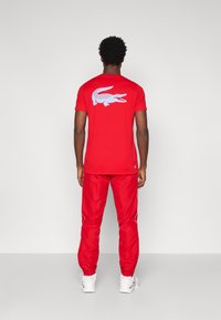 Rdeča majica s kratkimi rokavi z velikim belim grafičnim prikazom krokodila na hrbtni strani, v kombinaciji z rdečimi hlačami in belimi supergami.