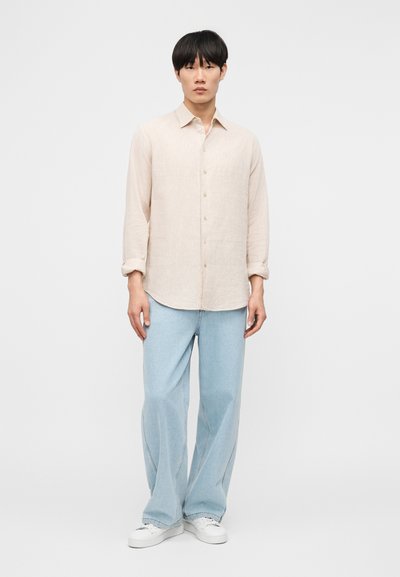 Άνδρας που φοράει μπεζ μακρυμάνικο πουκάμισο με κουμπιά, ανοιχτό μπλε τζιν παντελόνι με φαρδιά μπατζάκια και λευκά αθλητικά παπούτσια, στέκεται μπροστά σε απλό φόντο.