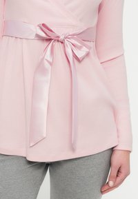 Top rosa con textura acanalada y mangas largas, con un lazo de satén en la cintura y dobladillo redondeado. Combinado con leggings de tela gris.