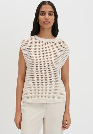 Femme aux longs cheveux foncés portant un pull sans manches en maille ouverte de couleur crème et un pantalon beige clair sur un fond uni.