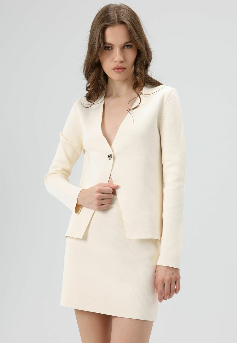 Costume deux pièces couleur crème comprenant un blazer à col en V profond avec manches longues, une fermeture à un bouton, et une mini-jupe assortie.