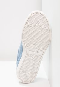 Semelle de chaussure blanche avec motif de bande de roulement texturé et nom de la marque "DIESEL" au centre, inclinée contre un mur d’angle blanc.