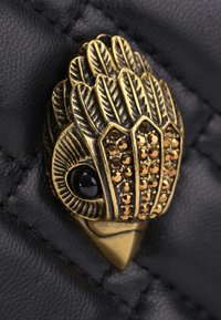 Decorazione a forma di uccello in metallo con dettagli di piume, adornata con strass marroni e un occhio in pietra nera, su uno sfondo di pelle nera testurizzata.