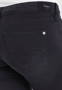 Black denim jeans with a back pocket featuring a curved stitching detail and metal rivet. Logo tag on waistband. Smooth texture.