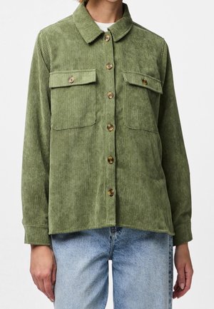 Groen corduroy overhemd met knopen en twee borstzakken, gedragen over een wit shirt en gecombineerd met lichtblauwe spijkerbroek.