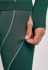 Leggings de sport extensibles verts avec une surface texturée et des coutures blanches en accent. Une main tire sur le tissu près de la taille.