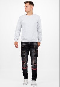 Sweatshirt gris à manches longues, associé à un jean noir avec plusieurs poches zippées et des accents rouges. Le modèle porte des baskets blanches.