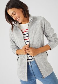 Giacca grigia strutturata con zip e colletto alto. Presenta tasche laterali e una vestibilità comoda, indossata sopra una maglietta a righe e jeans blu.