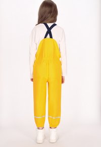 Dieťa v žltých nepremokavých overalov s čiernymi popruhmi, bielej dlhé tričko a biele čižmy, stojí otočené chrbtom proti jednoliatemu pozadiu.