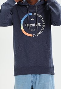Sudadera con capucha azul marino con un bolsillo canguro, que presenta un logo circular de Quiksilver en varios colores. Textura suave, cordones en la capucha.