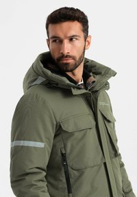 Giacca isolante verde oliva con cappuccio, multiple tasche frontali, dettagli riflettenti argento sulle maniche e finitura in tessuto testurizzato.