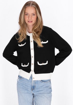 Kobieta o długich blond włosach, ubrana w czarny kardigan z białym obszyciem i guzikami, w połączeniu z jasnoniebieskimi dżinsami, stojąca z rękami na biodrach.