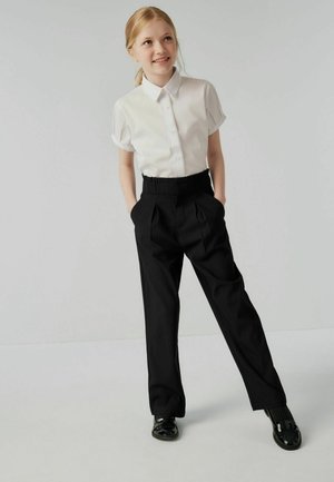 Chemise blanche à manches courtes avec des boutons et pantalon noir taille haute. Le pantalon présente des plis et des poches latérales. Chaussures noires avec une finition brillante.