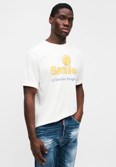 Λευκό βαμβακερό μπλουζάκι με γυρισμένα μανίκια, στολισμένο με κίτρινο γραφικό χαμογελαστού προσώπου και μπλε κείμενο: «Χαμογέλα, μπερδεύει τους ανθρώπους». Φορεμένο με σκισμένο μπλε τζιν.