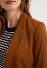 Rostfärgad blazer med en slät yta, med ett notch-krage. Under kanten syns en marinblå randig tröja med vita detaljer.