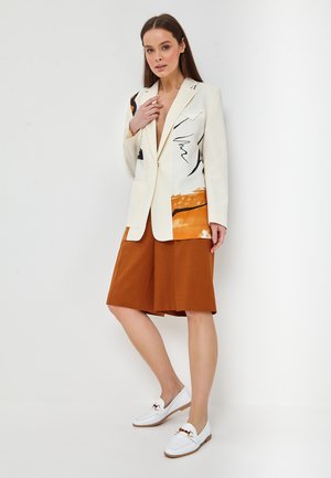 Kvinde iført en cremefarvet blazer med abstrakte sorte og orange mønstre, rustrøde shorts til knæet og hvide loafers, stående mod en hvid væg.