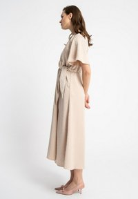 Jasno beżowa sukienka z krótkimi, falbaniastymi rękawami, wiązanym pasem w talii oraz zwiewnym krojem. Różowe szpilki z kokardkami dopełniają stylizację.