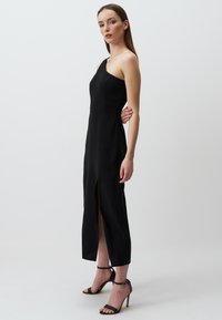 Abito nero con spallina unica realizzato in un tessuto liscio, caratterizzato da una fessura laterale e un design aderente. Indossato con tacchi a strappo neri.