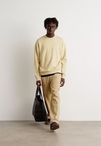 Beige mikina a světle hnědé kalhoty, které nosí osoba držící černou tašku. Outfit má jednoduchý design a uvolněný střih.