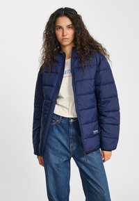 Marinblå vadderad jacka med quiltad design, ståkrage och dragkedja. Har en logga på den nedre framsidan och ribbade muddar.