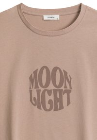 Jasnobrązowy t-shirt z okrągłym wzorem graficznym, na którym znajduje się napis "MOON LIGHT" w ciemniejszym odcieniu brązu. Miękka faktura materiału.