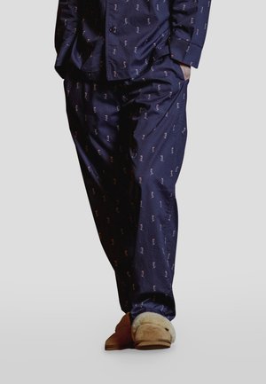 Persona con un conjunto de pijama azul marino con pequeños diseños estampados y zapatillas beige forradas de pelo, de pie con una mano en el bolsillo.