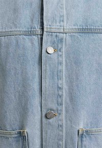 Jasnoniebieski denim z przeszyciem pionowym, dwoma metalowymi guzikami oraz częściowym widokiem przeszywanych kieszeni na dole.