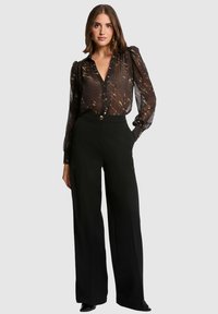 Blouse noire à boutons avec une texture transparente et de longues manches bouffantes, associée à un pantalon large noir taille haute avec des accents de boutons dorés.