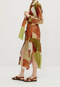 Robe longue multicolore avec des formes abstraites en orange, vert et beige, dotée d'un col et de fentes latérales. Associée à des sandales marron.