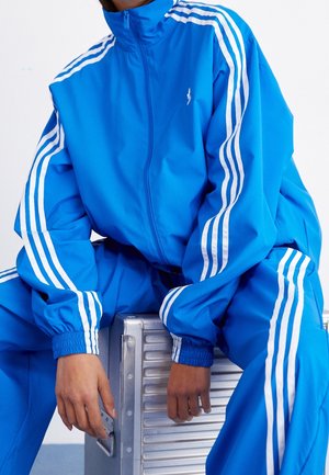 Personne en survêtement bleu avec des bandes blanches assise sur une valise métallique argentée, les mains reposant sur les poignées de la valise.