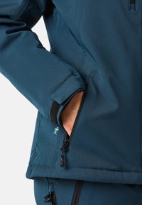 Jaket w kolorze teal z zamkniętą kieszenią boczną oraz akcentem w postaci flagi szwedzkiej. Teksturowany materiał z dopasowanym krojem i regulowanymi mankietami.