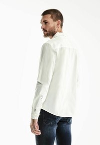 Bílá košile s dlouhým rukávem a límcem na knoflíky, vyrobená z texturované látky, zkombinovaná s tmavými džínami. Obsahuje manžety s detaily na knoflíky a švem.